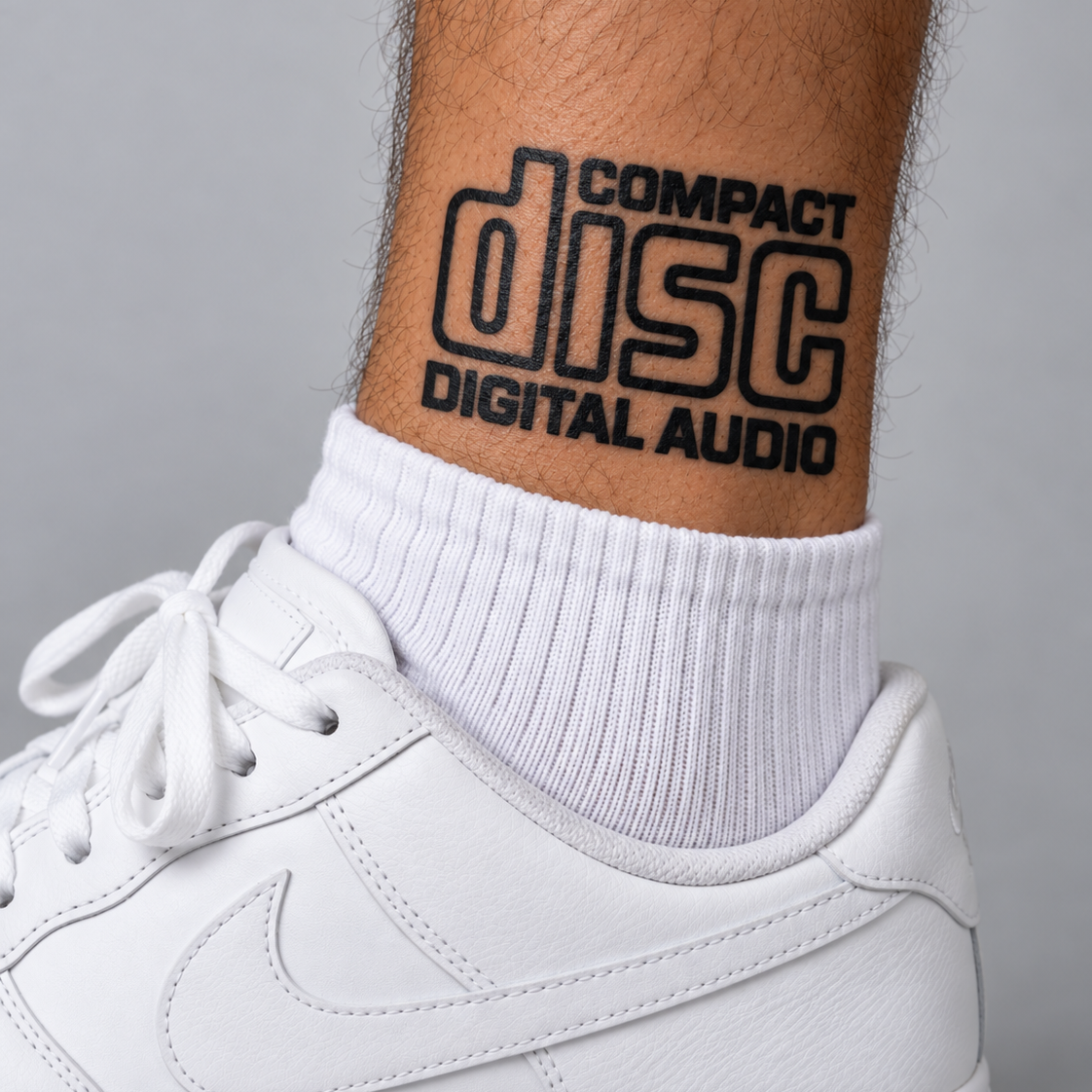A close-up of a person's calf wearing a white crew sock with a black 'Compact Disc Digital Audio' graphic, paired with a white Nike sneaker against a gray background.
